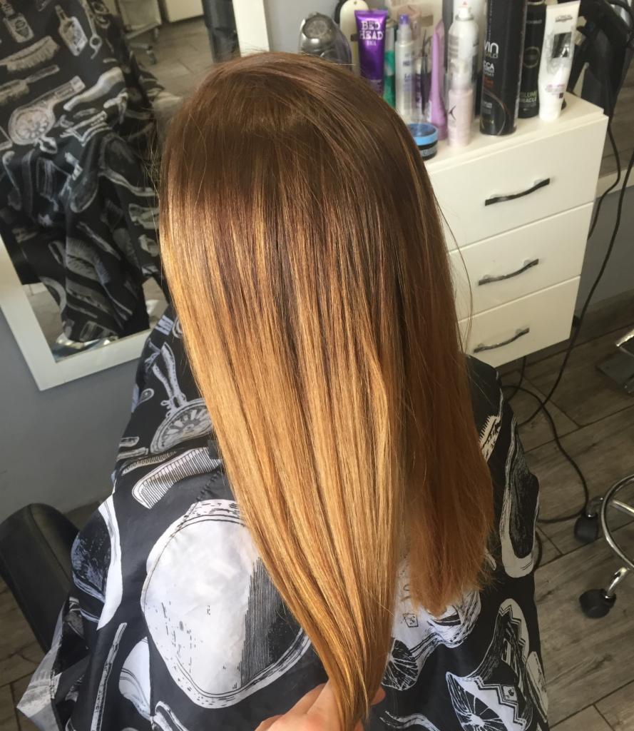 Świeżo pofarbowane, proste włosy w odcieniach miedzi i blondu, widoczne z tyłu, na tle salonu z kosmetykami i lustrem.