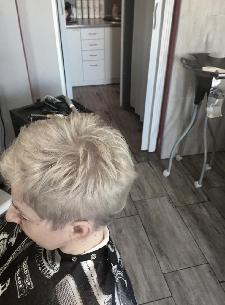 Krótkie, platynowe włosy po strzyżeniu, widoczne z góry, klientka w pelerynie ochronnej z motywem fryzjerskim, w tle fragment wnętrza salonu.