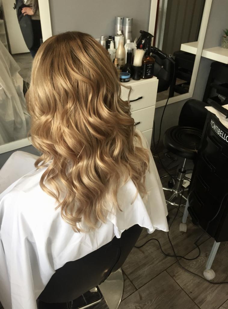 Średniej długości, pofalowane włosy w odcieniach blondu, widoczne z tyłu, po zabiegu stylizacji w salonie, z widocznym stanowiskiem pracy i akcesoriami.