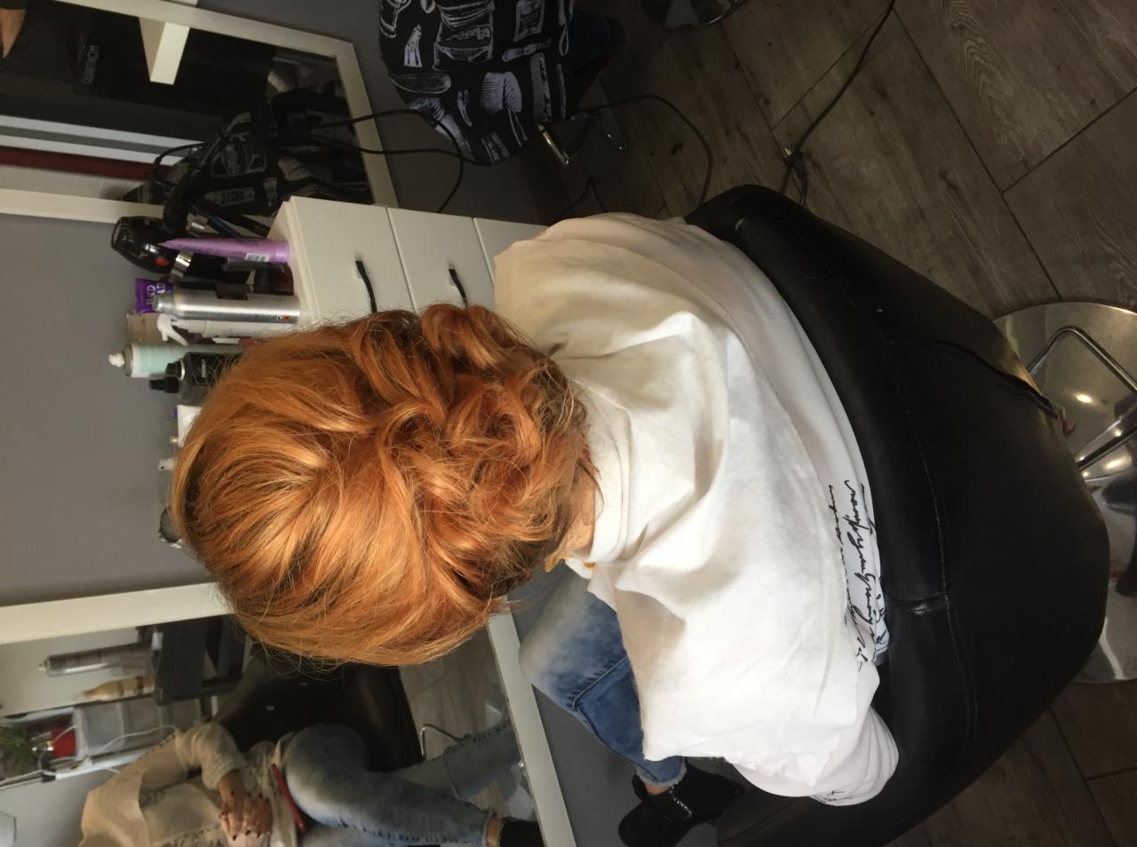 Widok z góry na głowę klientki z rudymi włosami upiętymi w salonie fryzjerskim, widoczny fotel fryzjerski i kosmetyki na półce.