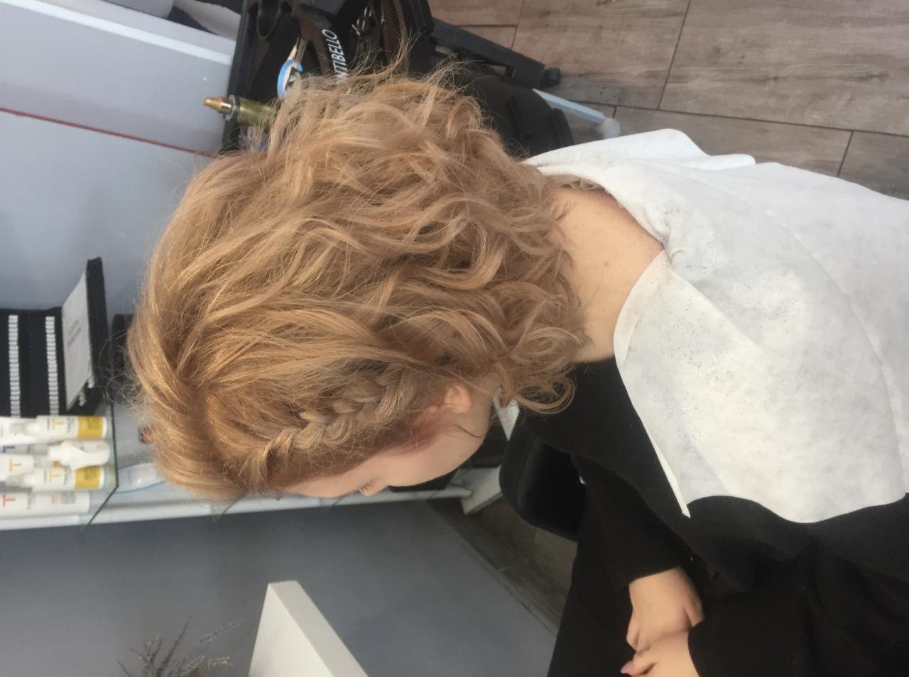 Kobieta z blond, kręconymi włosami i warkoczem przy skroni, zabezpieczona peleryną fryzjerską przed zabiegiem.