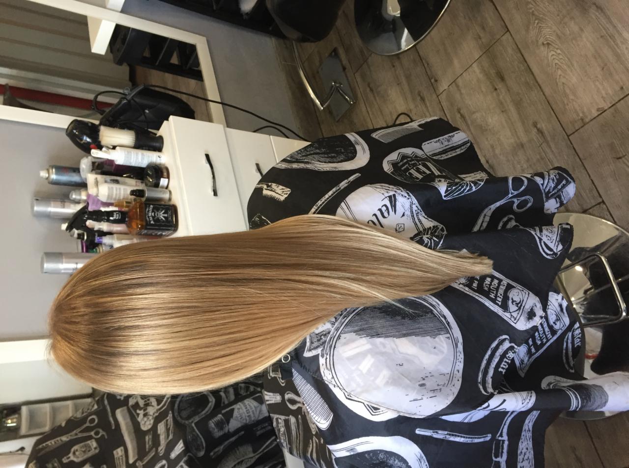 Świeżo ułożone, długie blond włosy na czarno-białym fryzjerskim fartuchu z wzorami narzędzi, w tle biała szafka z kosmetykami i lustro.