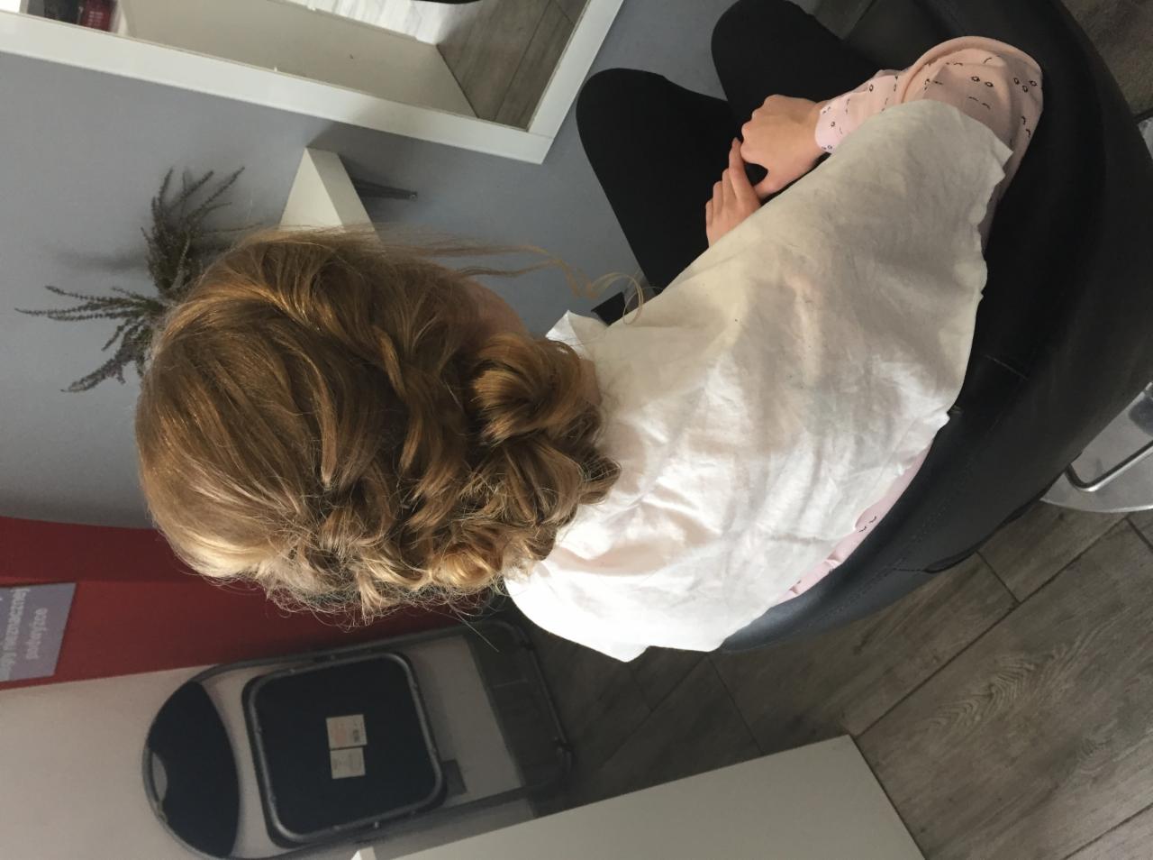 Upięcie włosów z tyłu głowy klientki siedzącej w fotelu fryzjerskim, włosy w odcieniach blondu, widoczne splecione pasma i upięcie w kok.