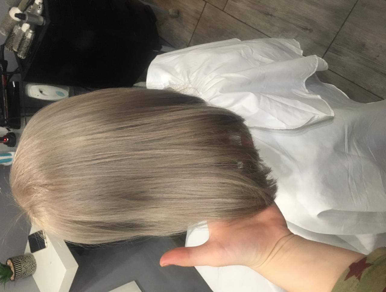 Świeżo ostrzyżone, gładkie, popielate włosy o długości do ramion, widoczne z tyłu głowy, trzymane przez dłoń na tle białego okrycia ochronnego.