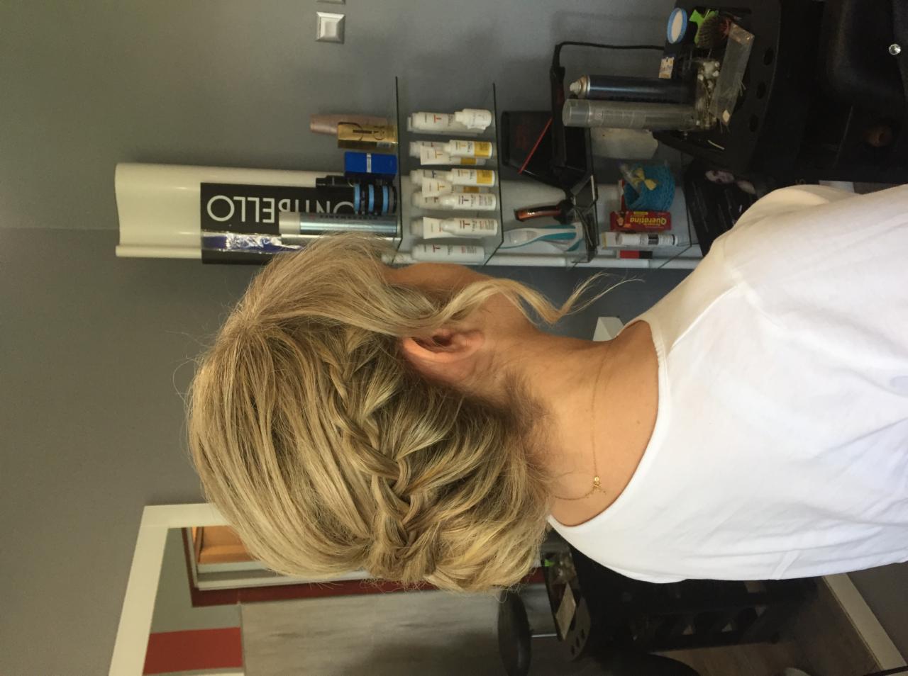 Blond włosy upięte w warkocz, widok z boku, w tle półka z kosmetykami i akcesoriami.