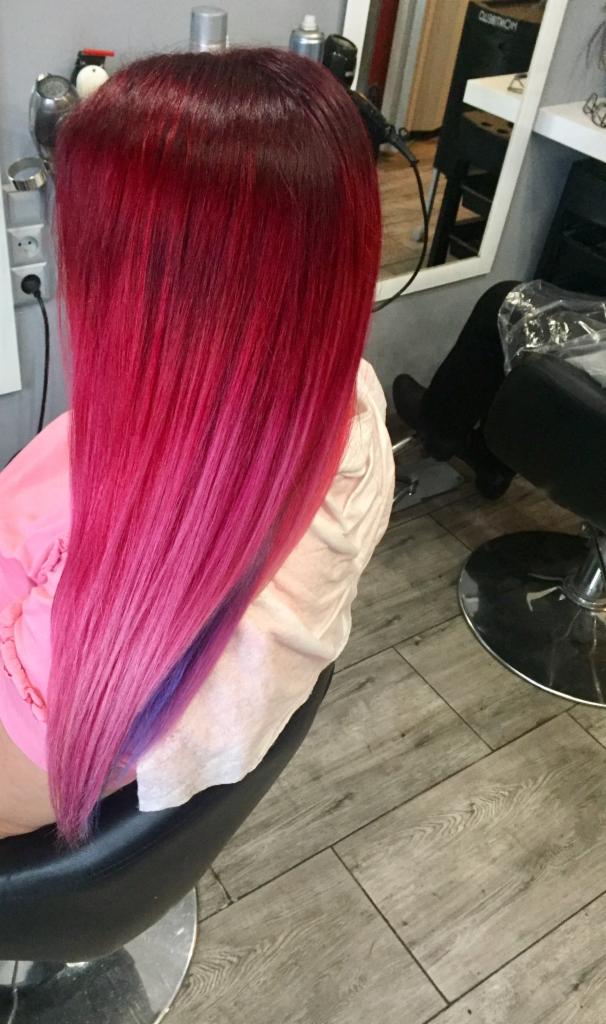 Długie włosy z koloryzacją ombre: od ciemnej czerwieni u nasady, przez intensywny róż, aż po fioletowe końcówki, widoczne na tle fryzjerskiego fotela i podłogi z paneli.