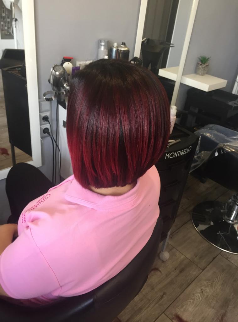 Krótkie, bordowo-czerwone włosy po koloryzacji i strzyżeniu w salonie fryzjerskim, widok z tyłu, na tle stanowiska fryzjerskiego z lustrem i akcesoriami.