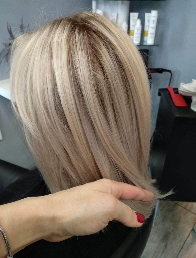 Blond włosy po koloryzacji z widocznymi odrostami w salonie fryzjerskim, uchwycone z boku, dłoń z czerwonym lakierem dotyka końcówek.