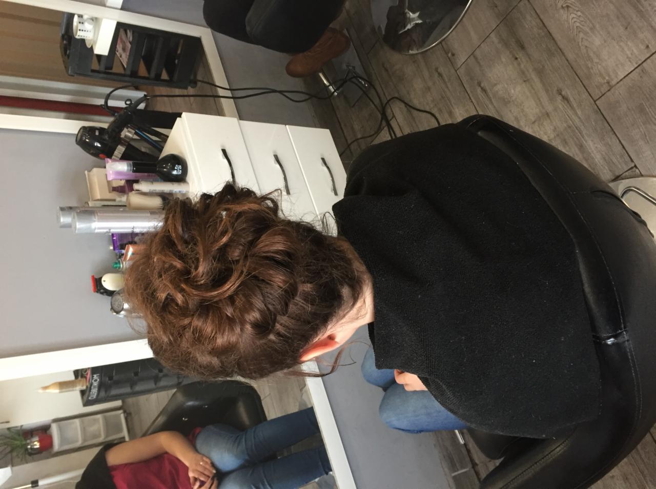Upięcie z brązowych włosów w salonie, widok z góry na tył głowy klientki w czarnym ubraniu siedzącej na fotelu, w tle biała szafka z kosmetykami i lustro.