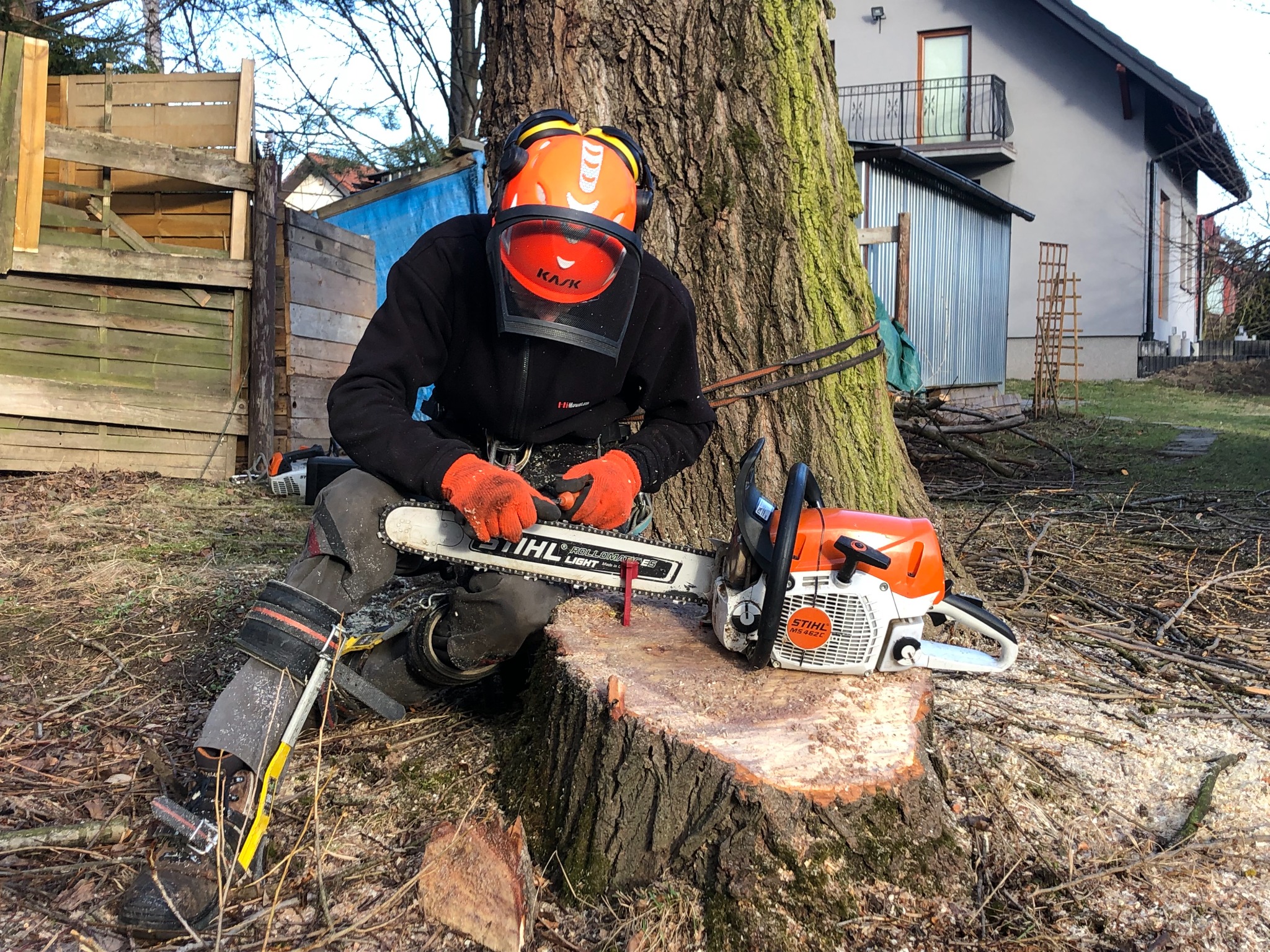 Arborysta w kasku i z pilarką Stihl MS 462 C siedzi na pniu drzewa po wycince, w tle budynek mieszkalny i drewniana konstrukcja.
