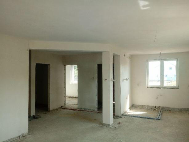 Surowe wnętrze domu w trakcie tynkowania maszynowego, widoczne ślady po tynku na ścianach i podłodze, okno z widokiem na budynek w tle.