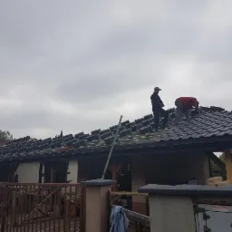 Dwóch dekarzy układa ciemną dachówkę na dachu domu jednorodzinnego w pochmurny dzień. Część dachu już pokryta, w tle widoczne przygotowane stosy dachówek.