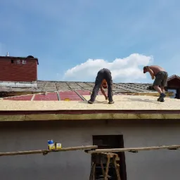 Dwóch mężczyzn bez koszulek układa płyty OSB na dachu domu jednorodzinnego w słoneczny dzień, widoczny komin z cegły i fragment starego pokrycia dachowego.