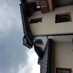 Fragment elewacji budynku z ciemną dachówką, rynnami i fragmentem kontenera na gruz na tle pochmurnego nieba.