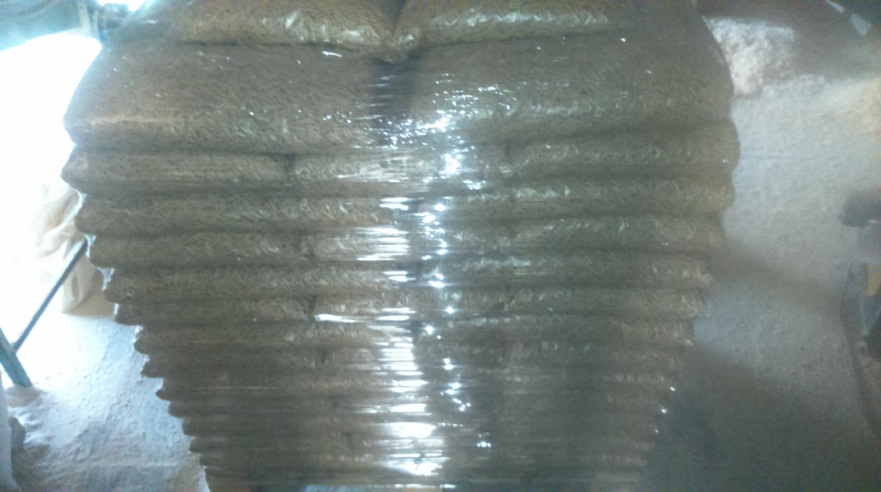 Stos worków z pelletem drzewnym, owiniętych folią, w magazynie, widoczne drobne jasne trociny na podłodze i słabe oświetlenie.