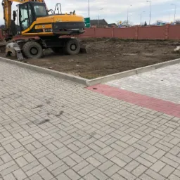 Żółta koparka kołowa JCB na placu budowy, obok ułożona szara kostka brukowa z czerwonym pasem, w tle przygotowany teren i ceglany mur.