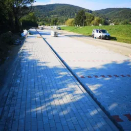 Długa, nowa droga z szarej kostki brukowej z czerwonymi akcentami, w tle góry i samochód dostawczy.