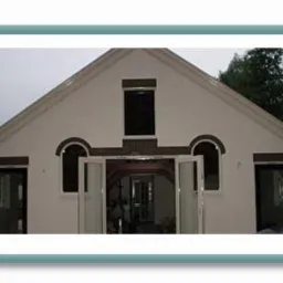 Wyremontowana fasada domu z białym tynkiem, ciemnymi oknami i otwartymi, przeszklonymi drzwiami balkonowymi.