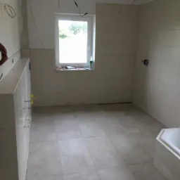 Wnętrze łazienki w trakcie kładzenia płytek ściennych i podłogowych, widoczne wystające przewody elektryczne i niezabudowana wanna narożna.