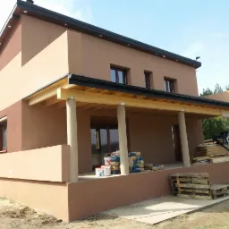 Dwukondygnacyjny dom jednorodzinny z jasnobrązową elewacją, drewnianą konstrukcją zadaszenia tarasu wspartą na kolumnach, workami z zaprawą budowlaną i paletami na tarasie, częściowo ocieplony...