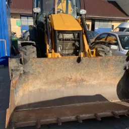 Żółta koparko-ładowarka z widocznymi śladami użytkowania na łyżce, zaparkowana na brukowanym terenie przed budynkiem z czerwonym dachem i zaparkowanymi samochodami.