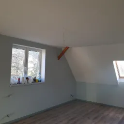 Pomalowane poddasze z drewnianymi belkami, oknem dachowym i oknem na ścianie, przygotowane do dalszych prac wykończeniowych.