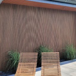 SWIAT DESKI - Nowoczesna elewacja z drewnianych paneli pionowych, uzupełniona kamienną ścianą i dwoma drewnianymi leżakami na szarym bruku. Minimalistyczny design.