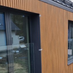 SWIAT DESKI - Elewacja budynku wykończona panelami w odcieniach brązu, z widocznymi oknami w ciemnych ramach i szklaną balustradą. Widok z zewnątrz, w tle drzewa.