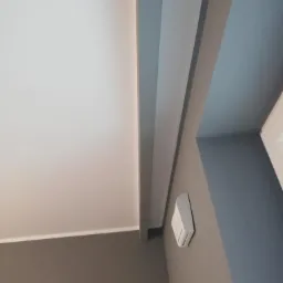 Pomieszczenie z pomalowanymi ścianami w odcieniach szarości i bieli, widoczne listwy dekoracyjne pod sufitem i czujnik na ścianie.