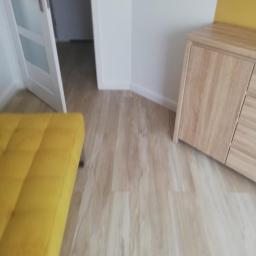 UsługiRemontowoBudowlane Tomasz Graczyk - Jasna podłoga z paneli imitujących drewno, widoczny fragment żółtej sofy, białe drzwi z przeszkleniami i fragment drewnianej komody w rogu pokoju.