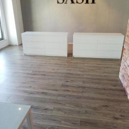 UsługiRemontowoBudowlane Tomasz Graczyk - Ułożona podłoga z paneli imitujących drewno w nowym biurze, widoczne dwie białe komody i fragment ściany z cegły.