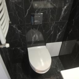UsługiRemontowoBudowlane Tomasz Graczyk - Nowoczesna toaleta wisząca z chromowanym przyciskiem spłukiwania, otoczona czarnymi płytkami z białym marmurkowym wzorem i białym grzejnikiem drabinkowym po lewej stronie.