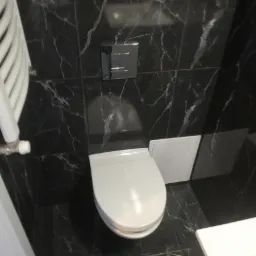 Nowoczesna toaleta wisząca z chromowanym przyciskiem spłukiwania, otoczona czarnymi płytkami z białym marmurkowym wzorem i białym grzejnikiem drabinkowym po lewej stronie.