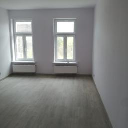 UsługiRemontowoBudowlane Tomasz Graczyk - Pomalowane na szaro wnętrze pokoju z dwoma oknami i ułożonymi panelami podłogowymi w odcieniach szarości, widoczne grzejniki pod oknami, brak listew przypodłogowych.