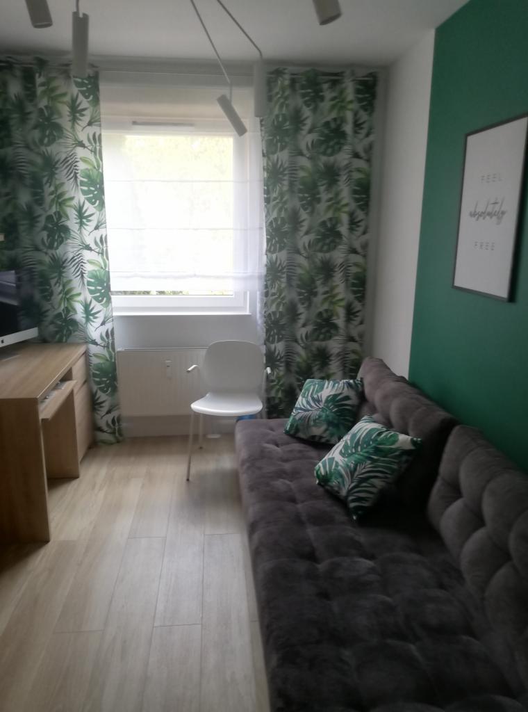 Pokój z szarą sofą, poduszkami w tropikalne liście, zasłonami w ten sam wzór, białym krzesłem i zieloną ścianą z plakatem.