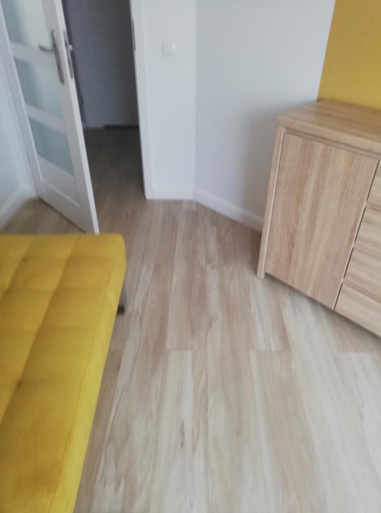Jasna podłoga z paneli imitujących drewno, widoczny fragment żółtej sofy, białe drzwi z przeszkleniami i fragment drewnianej komody w rogu pokoju.