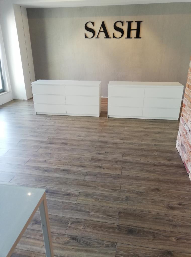 Ułożona podłoga z paneli imitujących drewno w nowym biurze, widoczne dwie białe komody i fragment ściany z cegły.