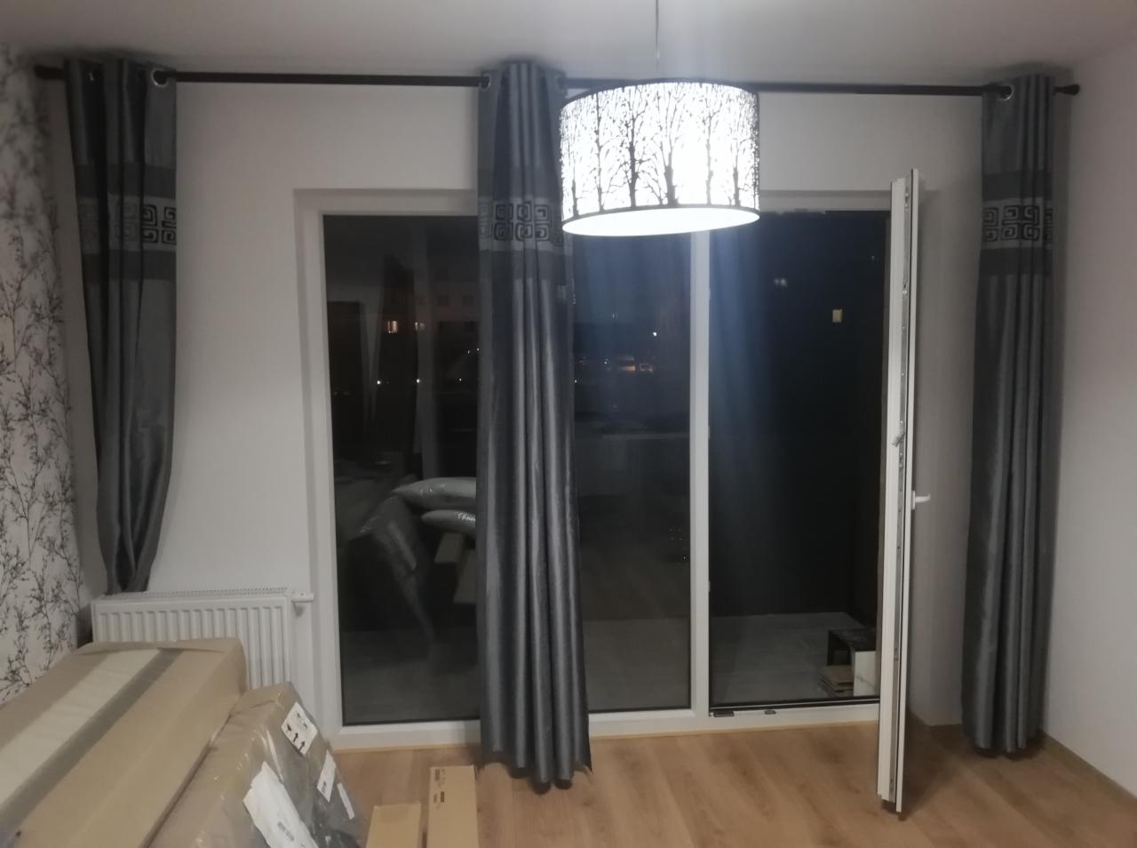 Pokój z szarymi zasłonami z geometrycznym wzorem, lampą sufitową z motywem drzew i podłogą w odcieniach drewna, z widocznymi paczkami do rozpakowania.