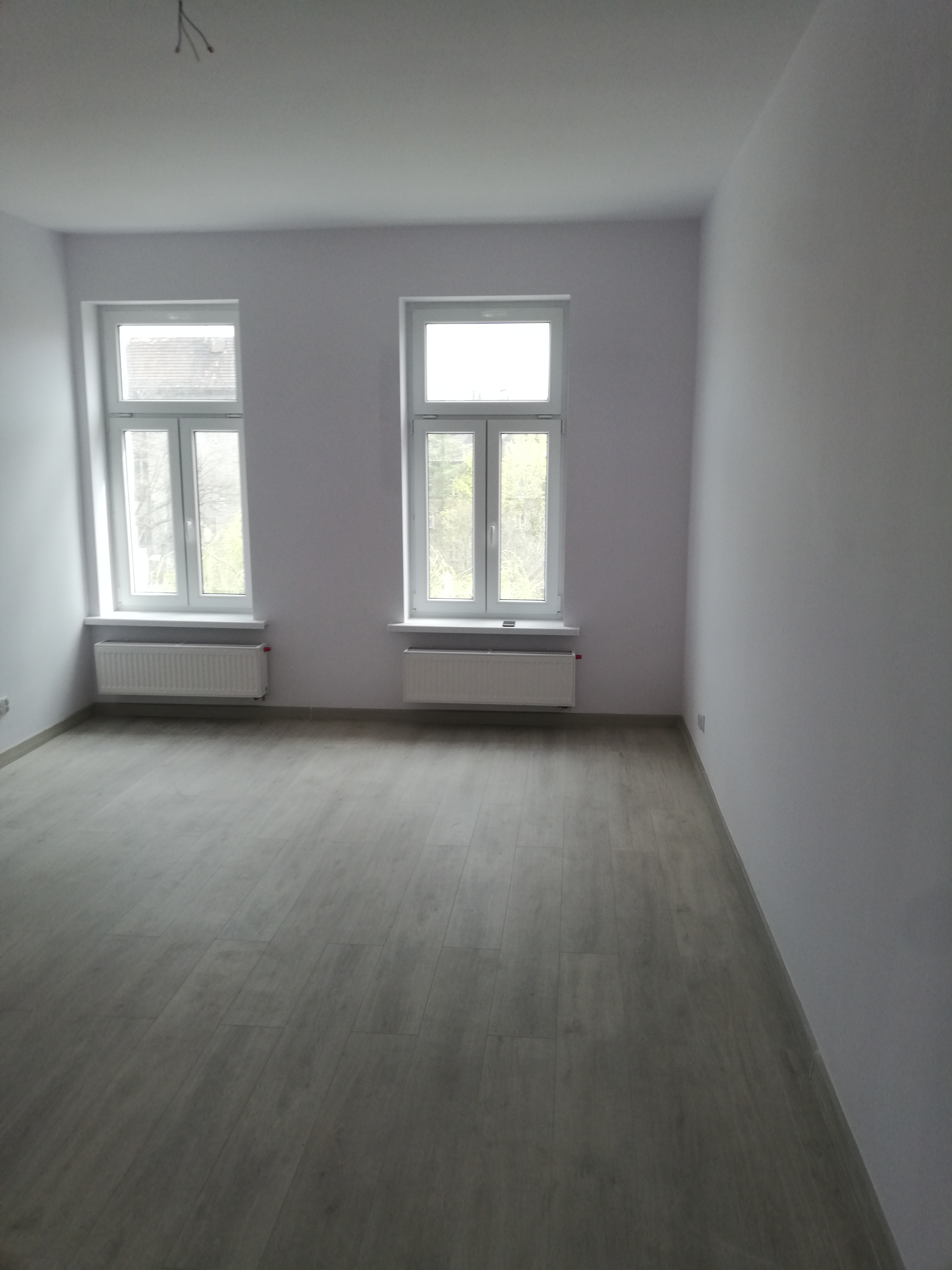 Pomalowane na szaro wnętrze pokoju z dwoma oknami i ułożonymi panelami podłogowymi w odcieniach szarości, widoczne grzejniki pod oknami, brak listew przypodłogowych.