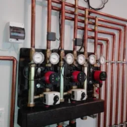 Rozdzielacz ogrzewania podłogowego z rurami miedzianymi, manometrami, pompami obiegowymi i sterownikami, zamontowany na ścianie w pomieszczeniu technicznym.