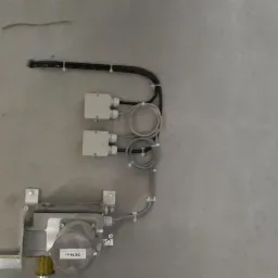 Szara ściana z zamontowanym metalowym urządzeniem z naklejką '50Nm', dwoma białymi puszkami elektrycznymi i przewodami przymocowanymi do ściany za pomocą uchwytów kablowych.