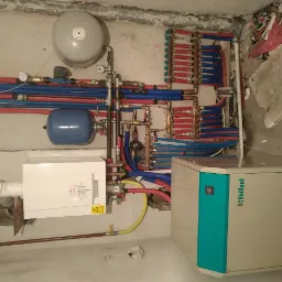 Kompleksowa instalacja grzewcza w kotłowni: biały piec Vaillant, rozdzielacz ogrzewania podłogowego z rurami w kolorach niebieskim i czerwonym, dwa zbiorniki wyrównawcze, rury miedziane...