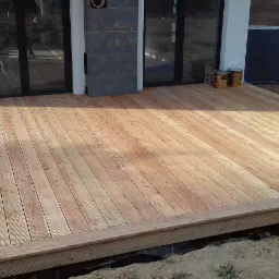 Nowo wybudowany taras z jasnego drewna, z widocznymi puszkami po impregnacie, przylegający do nowoczesnego domu z dużymi oknami tarasowymi.