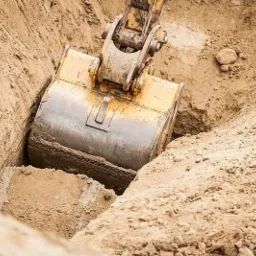 Żółta koparka zagłębia się w wąski wykop w piaszczystej ziemi, świeżo usypana pryzma piasku na pierwszym planie.