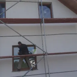 Pracownik na rusztowaniu przy białej ścianie domu, widoczne okna z brązowymi ramami i wiadro farby.