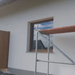 Świeżo otynkowana, biała elewacja domu jednorodzinnego z drewnianymi oknami i drzwiami, widoczne rusztowanie z drewnianą deską, odblask nieba w szybach okiennych.