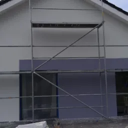 Dom jednorodzinny w trakcie malowania elewacji na dwa kolory: biały i fioletowy, widoczne rusztowania i zabezpieczone okna.