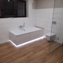 Nowoczesna łazienka z podświetlaną wanną, drewnopodobną podłogą i kabiną prysznicową ze szkła.