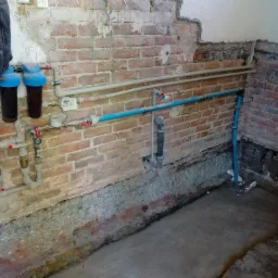 Fragment ściany z cegły z widocznymi rurami instalacji wodnej i grzewczej, filtry wody z niebieskimi pokrywami, manometr, oraz fragment podłogi w trakcie prac remontowych.