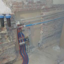 Edward Wiśniewski - Wnętrze w trakcie prac instalacyjnych: widoczny rozdzielacz z czerwono-niebieskimi rurami, filtry wody, oraz odsłonięta ceglana ściana z fragmentami tynku i przewodami elektrycznymi.